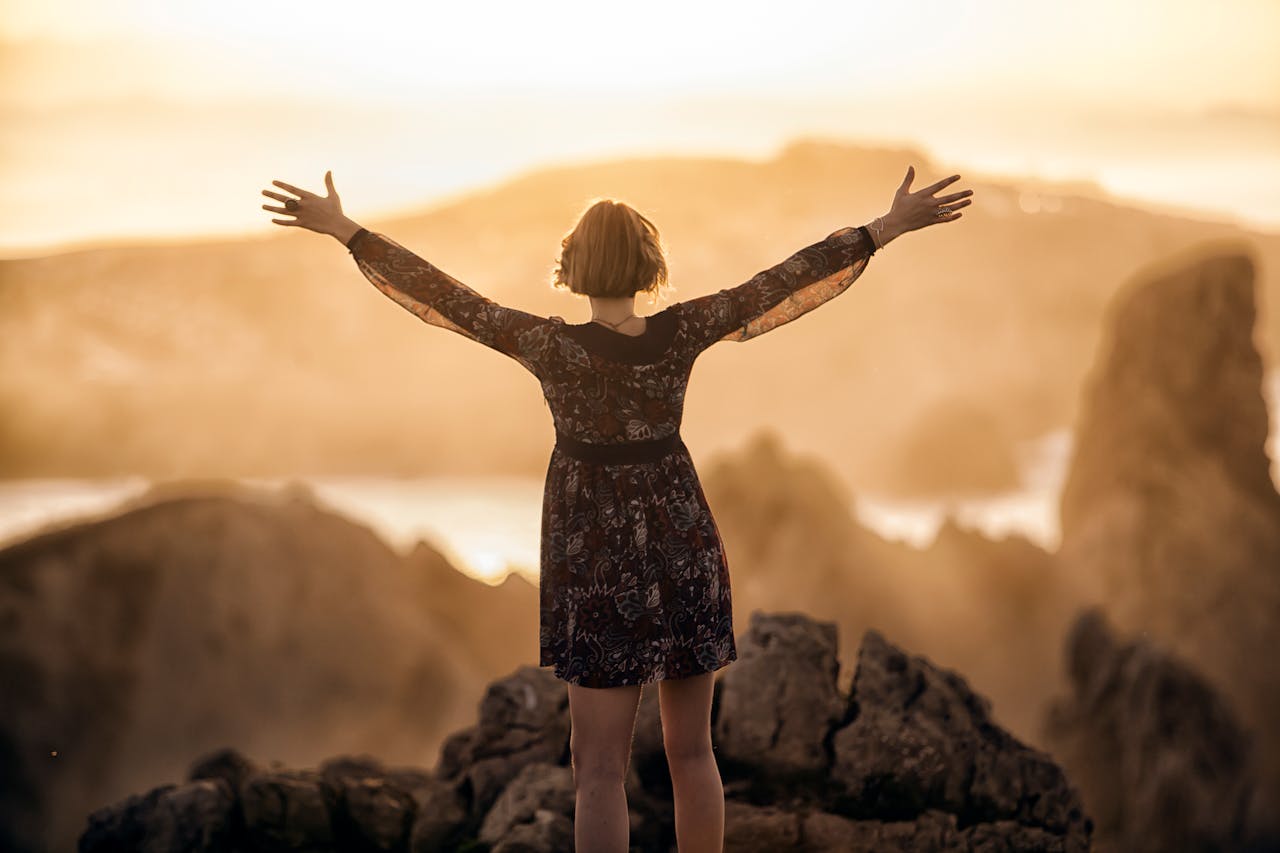A woman stands with open arms on a mountain at sunset, embracing nature's beauty.