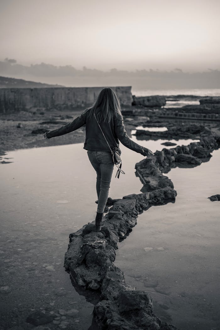 gallery-05 A woman walks along a rocky path by the water at twilight, exuding a sense of adventure and solitude.