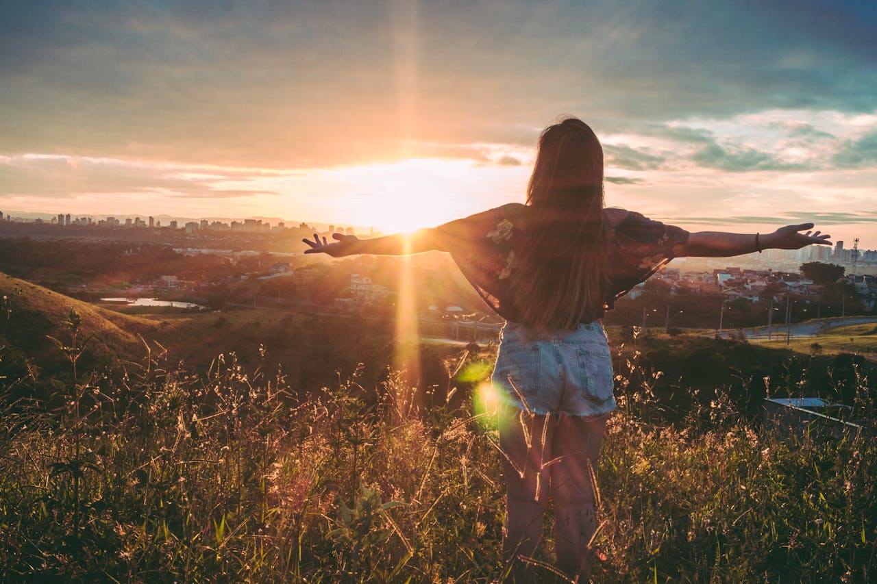 gallery-04 Woman with open arms enjoying sunset view over cityscape from a grassy hilltop.