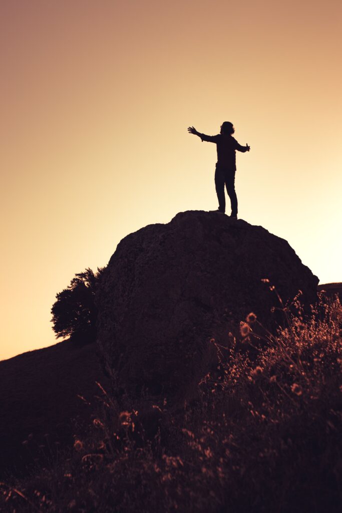 pexels photo 15386515 15386515 Silhouette of an individual with arms outstretched atop a rock against a yellow sunset sky in California.